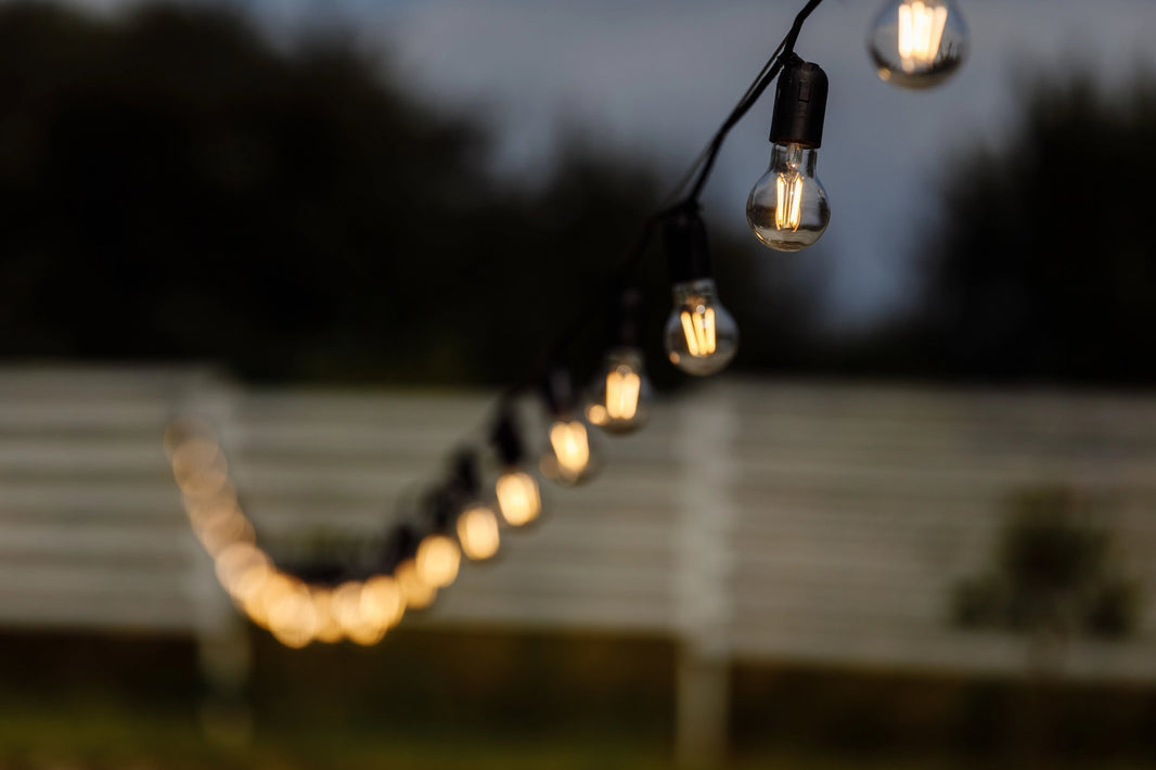 Closeup of a long string of incandescent globe-bulb string lights hanging outdoors at twilight, shining faintly.