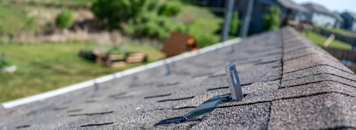 A closeup of an asphalt rooftop, on which a metal roof anchor is attached, and behind which a blurry, green residential neighborhood is visible.