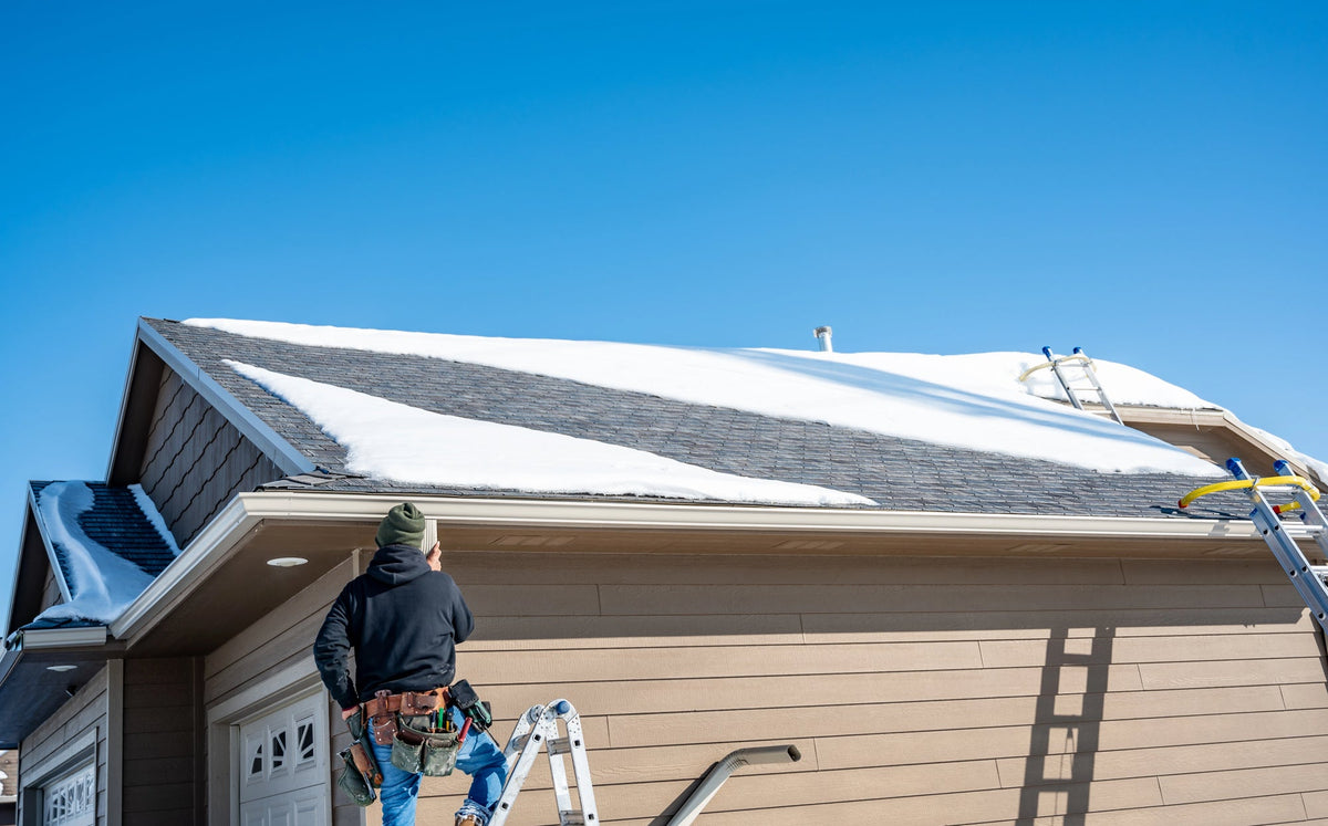 A contractor with a toolbelt examining the edge of a snow-covered rooftop while standing on a ladder.