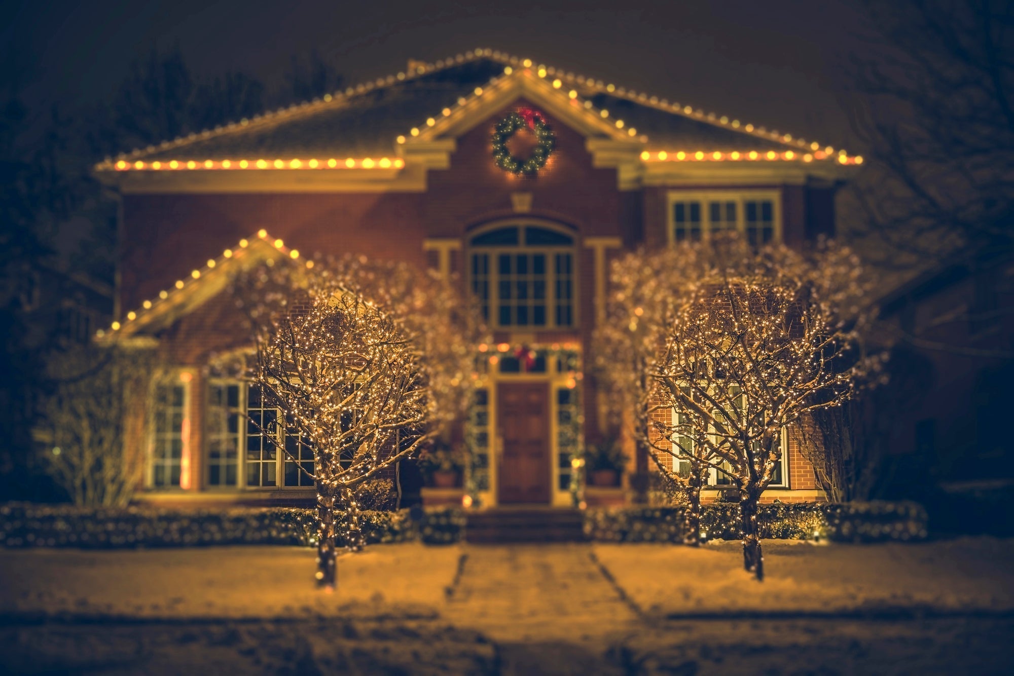 A warmly lit picture of a courtly brick building at night, covered in yellow Christmas lights and flanked by two barren trees in the snow, also covered in yellow lights.